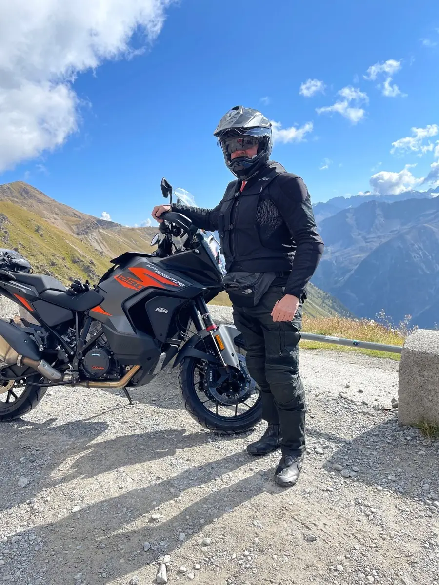 A rider in black adventure kit standing beside a grey and orange KTM Super Adventure on a gravel pull-off at the top of the Stelvio Pass, with autumn-gold slopes and blue sky behind