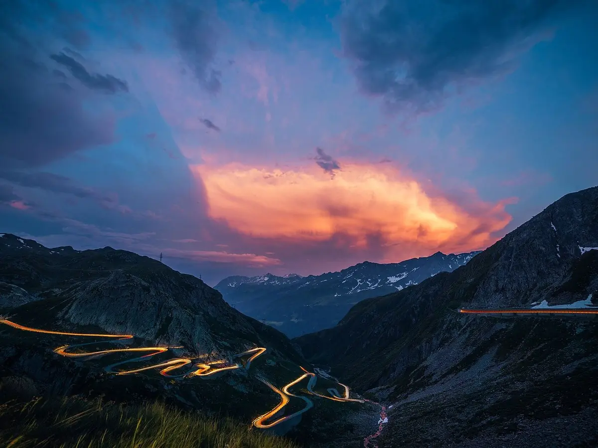 The cobbled hairpins of the Tremola old Gotthard road stacked up the mountainside above Airolo at dusk, headlights tracing the bends and a band of orange cloud over the valley